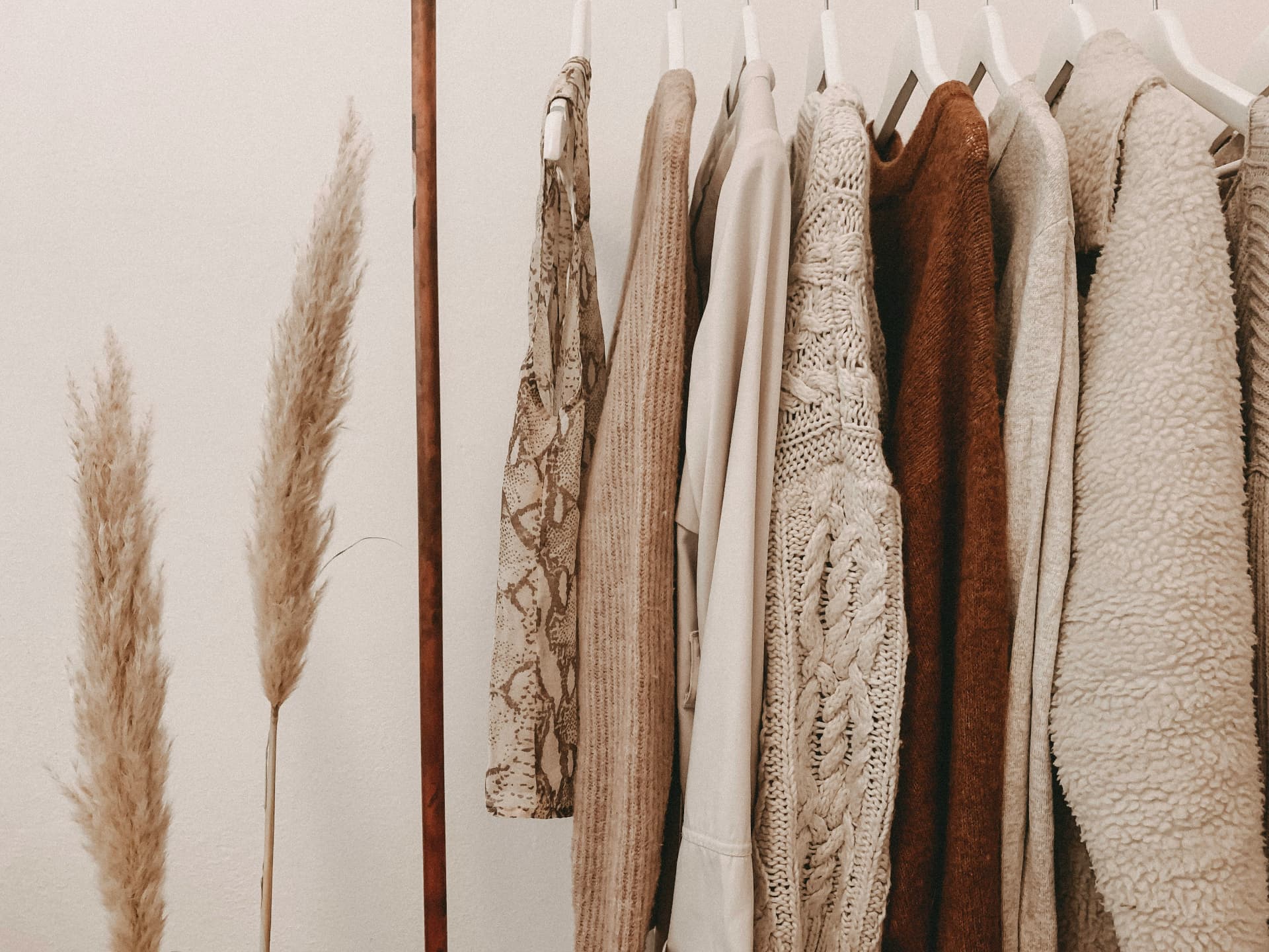 A neutral clothing rail of cream and camel knitwear beside dried pampas grass